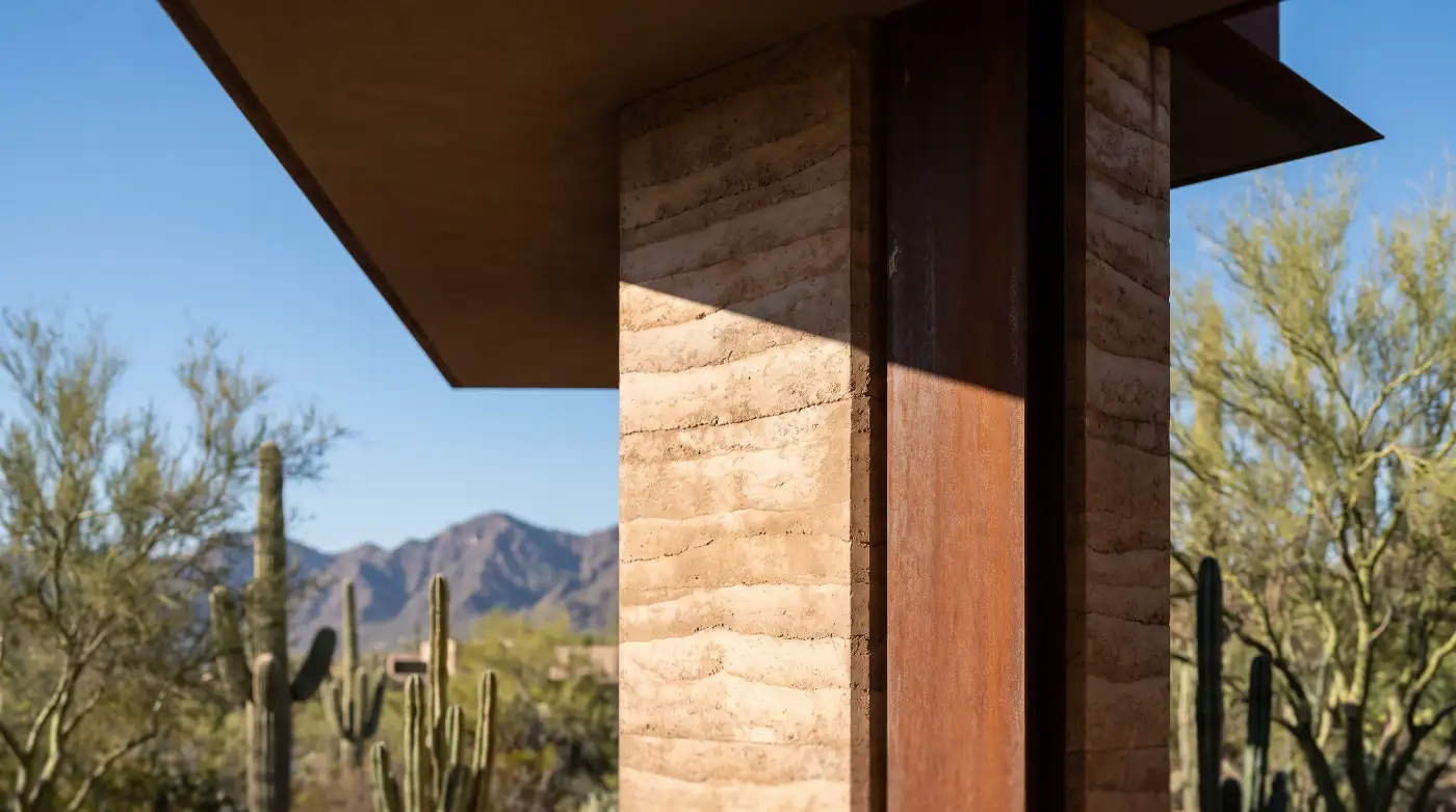 A close study of layered rammed earth meeting a weathered steel column under the hard shadow line of the overhang